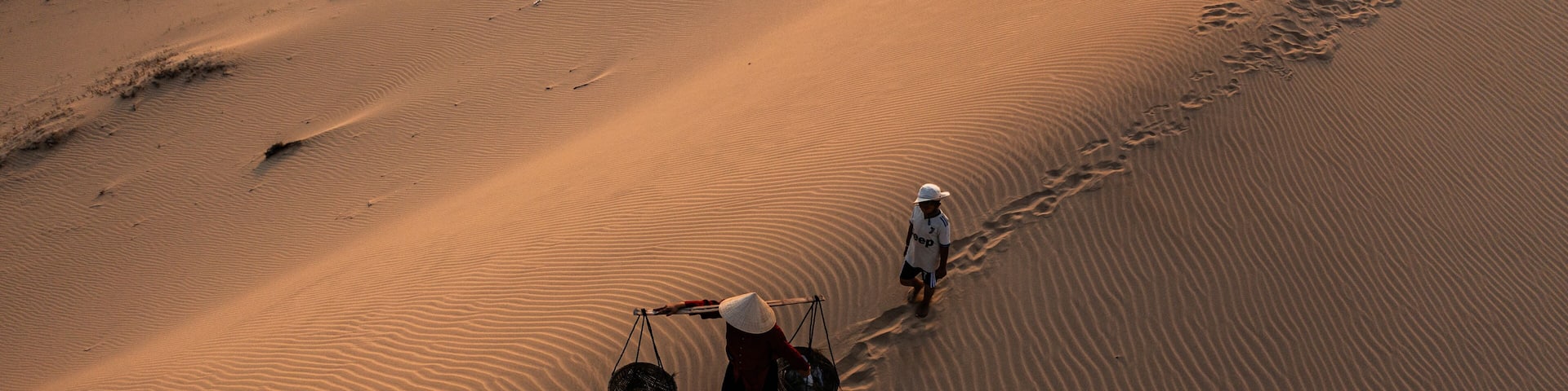 A vast desert landscape with rippled sand dunes and two individuals walking along a trail.