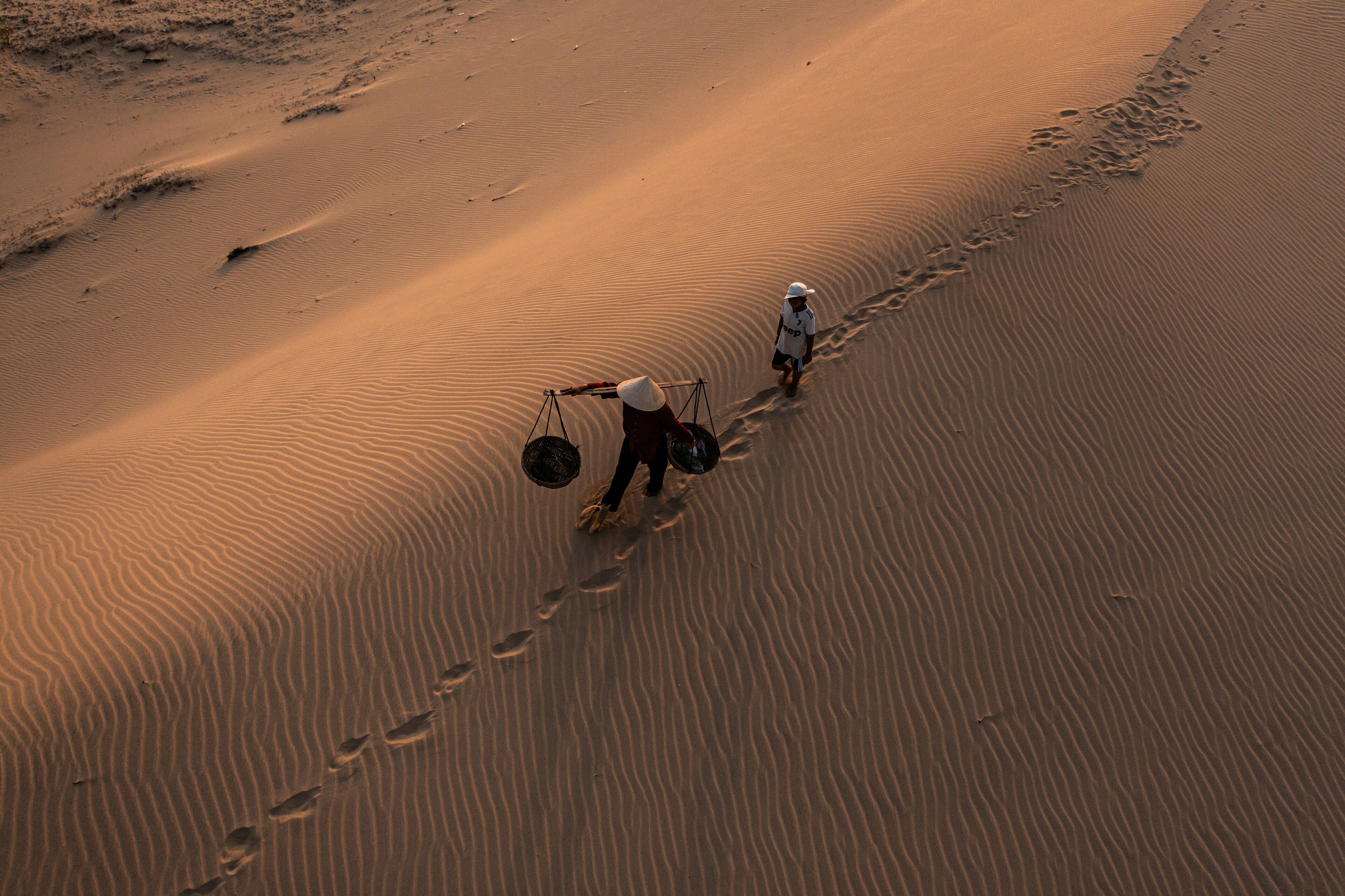 A vast desert landscape with rippled sand dunes and two individuals walking along a trail.