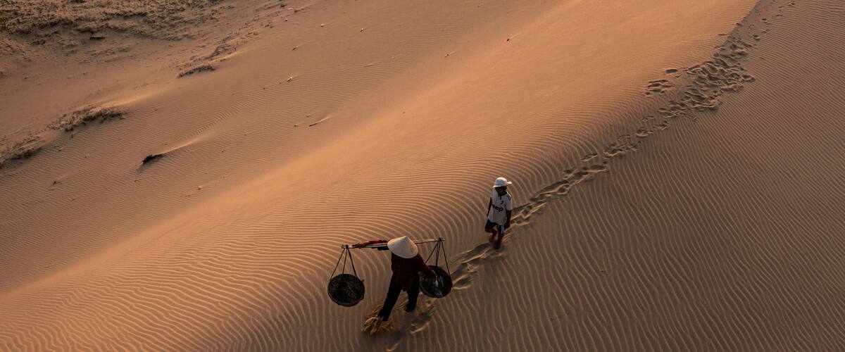 A vast desert landscape with rippled sand dunes and two individuals walking along a trail.