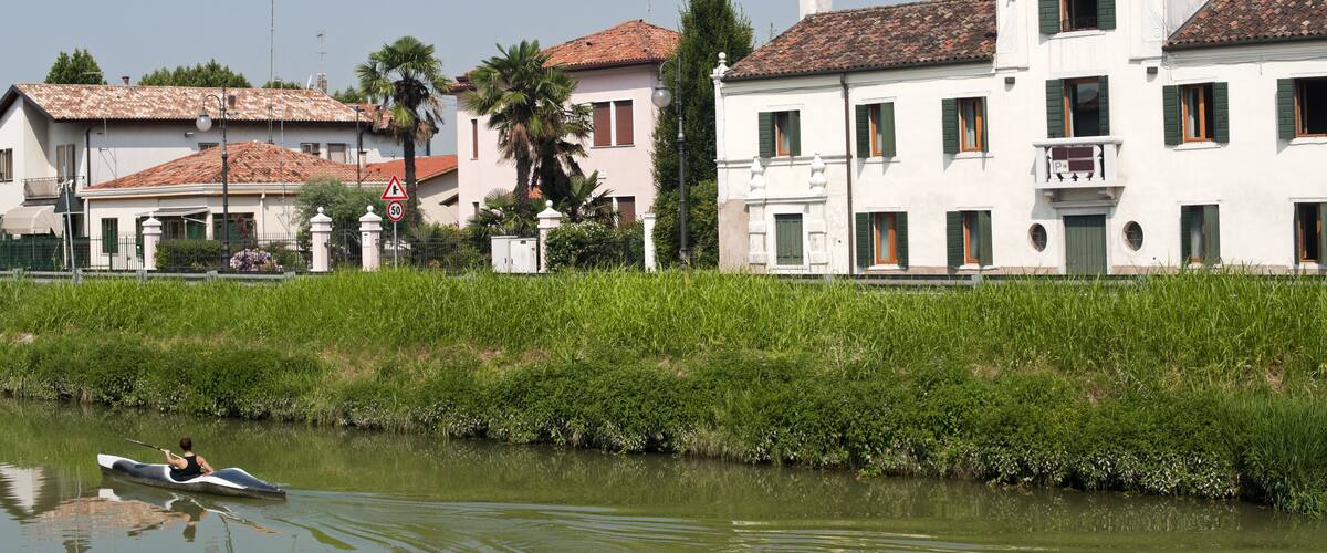 A calm river with a person rowing a boat, surrounded by lush greenery and traditional buildings with red-tiled roofs.