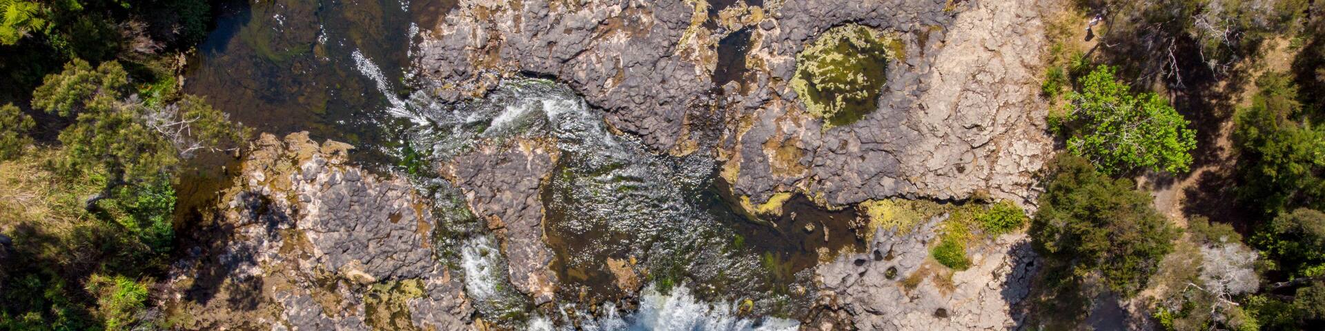 Aerial view of the Haruru Falls cascading over rugged, dark rocks into a serene, emerald pool surrounded by lush greenery, Haruru Falls, Bay of Islands, New Zealand.
