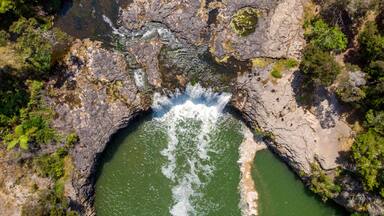 Aerial view of the Haruru Falls cascading over rugged, dark rocks into a serene, emerald pool surrounded by lush greenery, Haruru Falls, Bay of Islands, New Zealand.