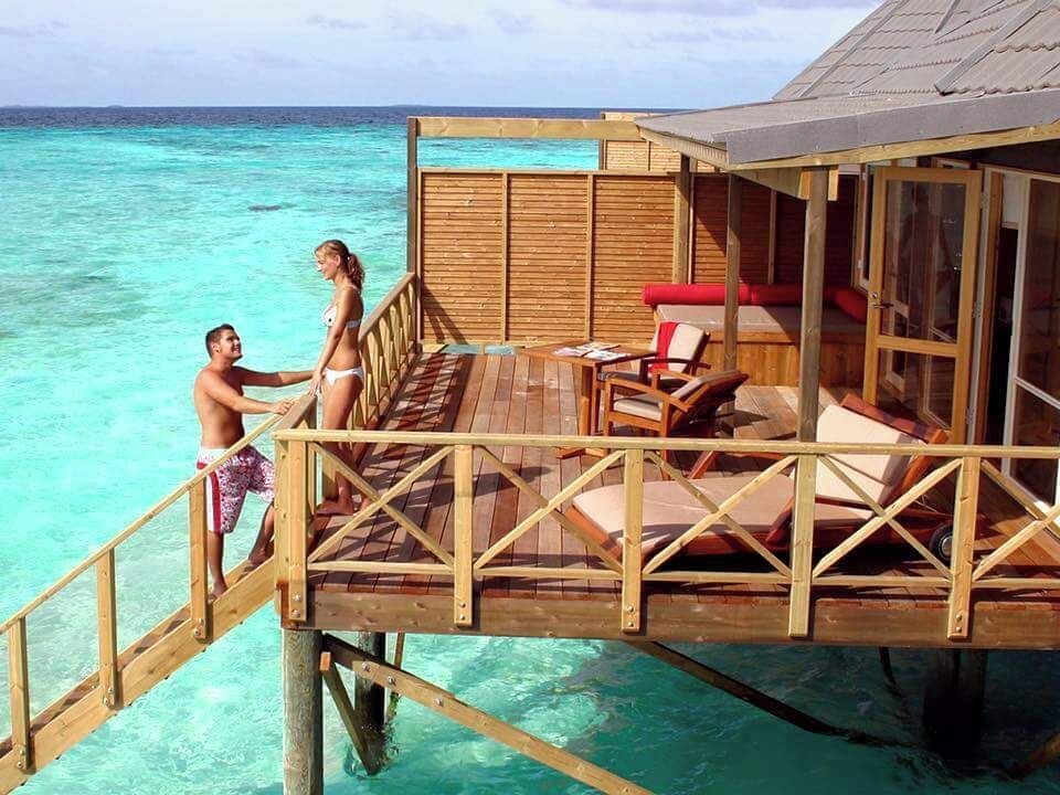 Um deck de madeira se estende sobre águas oceânicas azul-turquesa, com um casal em pé no deck e uma pequena estrutura com um assento acolchoado vermelho.