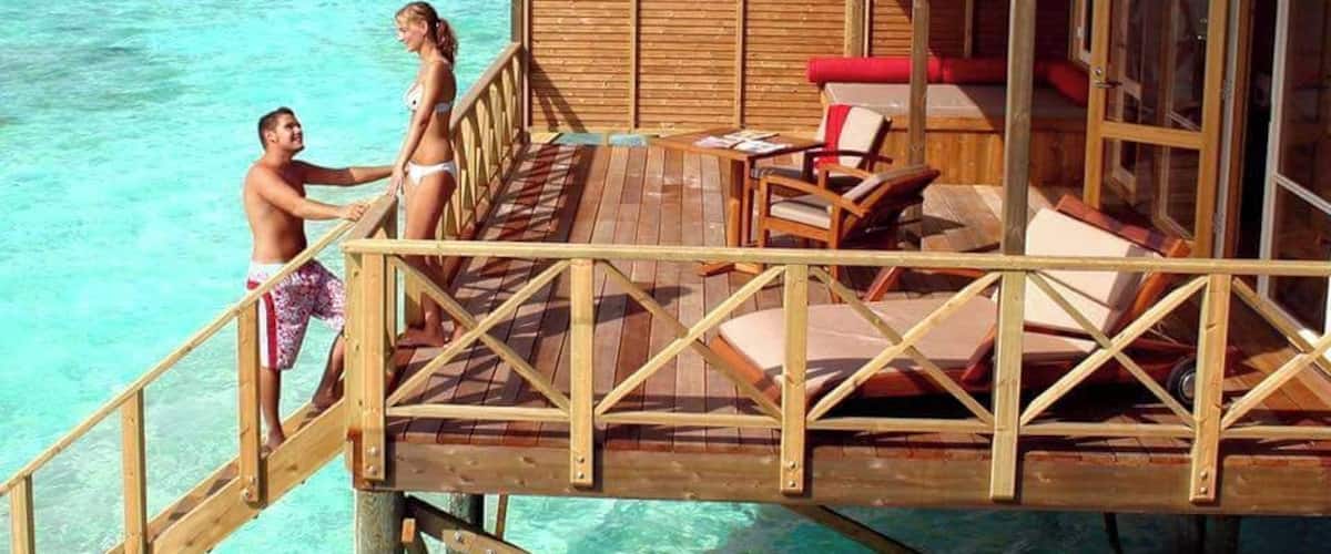 A wooden deck extending over turquoise ocean water, with a couple standing on the deck and a small structure with a red cushion.