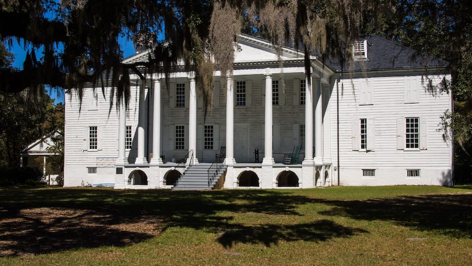 A large white building with columns and a porch, surrounded by trees and grass.