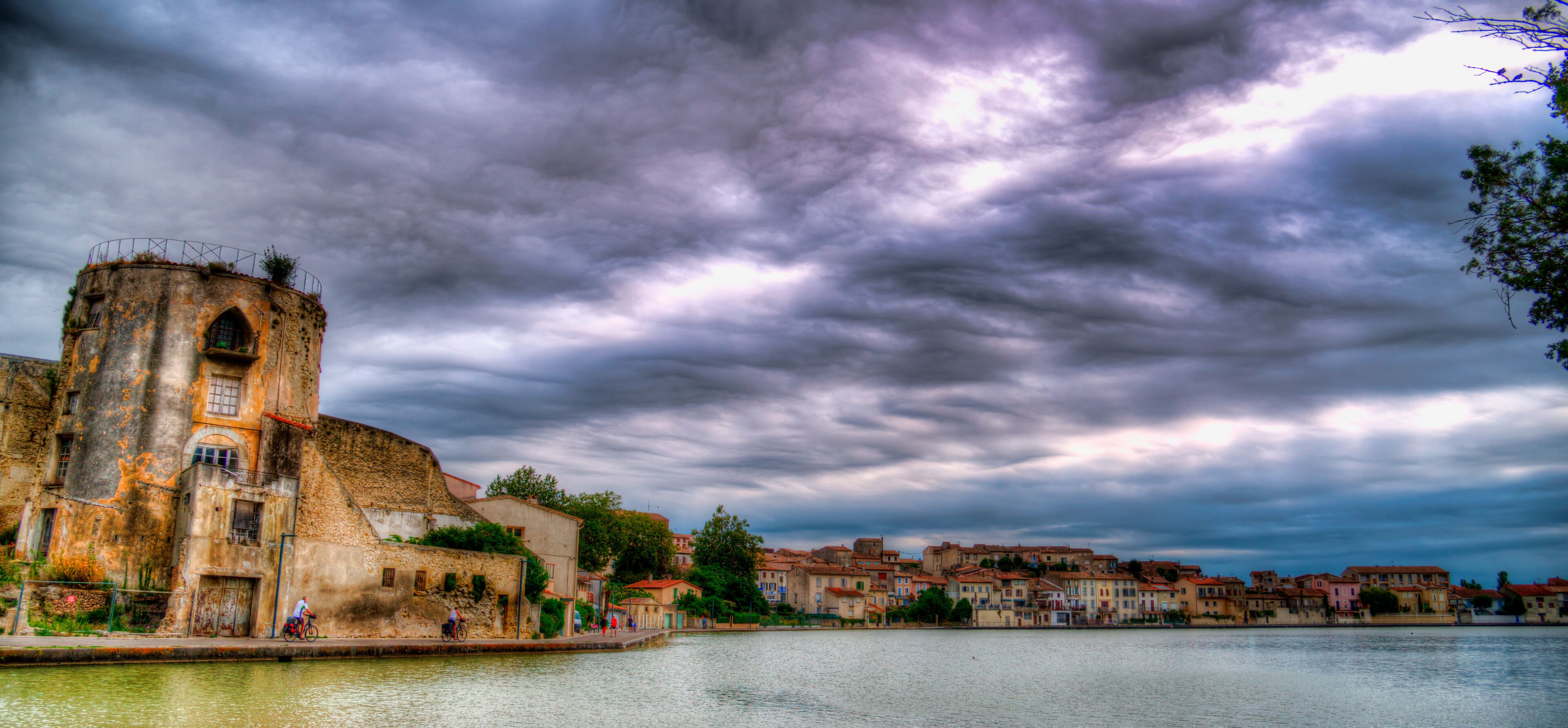 A historic building with a tower, a river, and a town with colorful buildings under a cloudy sky.