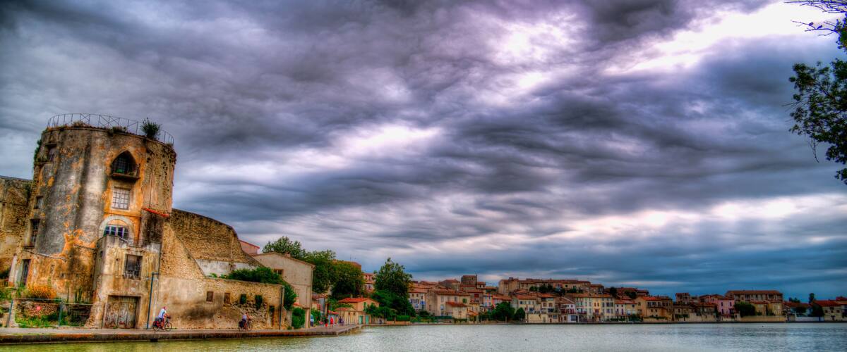 A historic building with a tower, a river, and a town with colorful buildings under a cloudy sky.