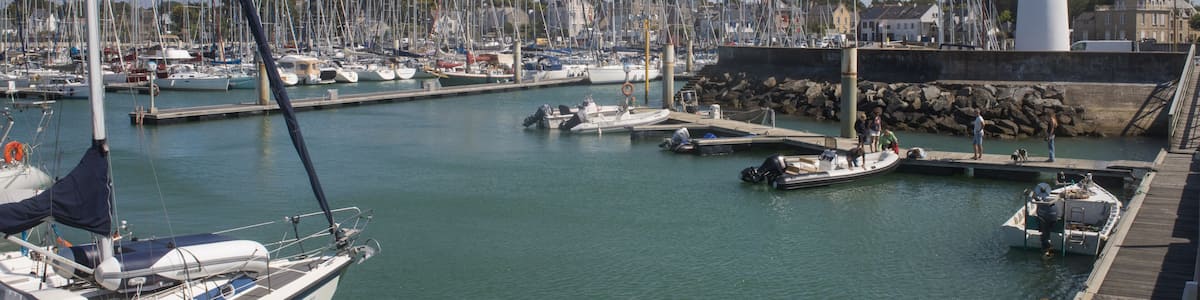 A marina with multiple boats docked, a lighthouse, and a rocky breakwater.