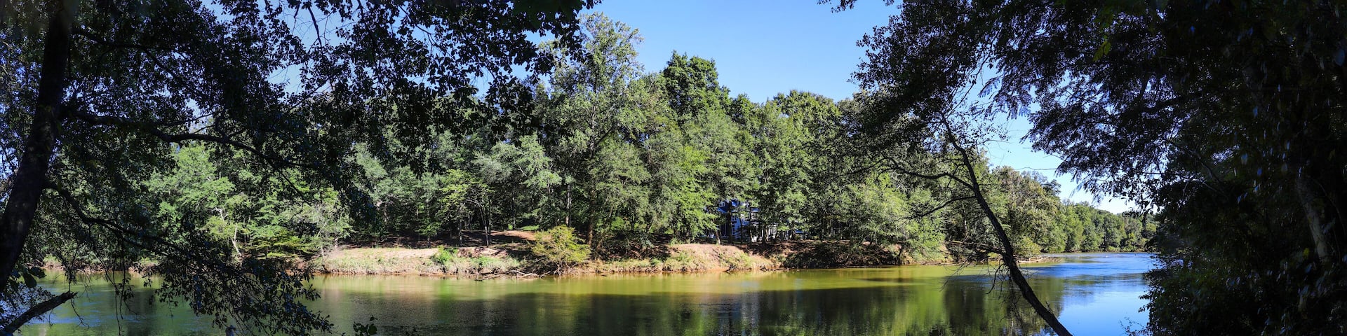 a gorgeous panoramic shot of the silky brown waters of the Chattahoochee river surrounded by lush green trees with blue sky at Jones Bridge Park Trail in Johns Creek Georgia USA
