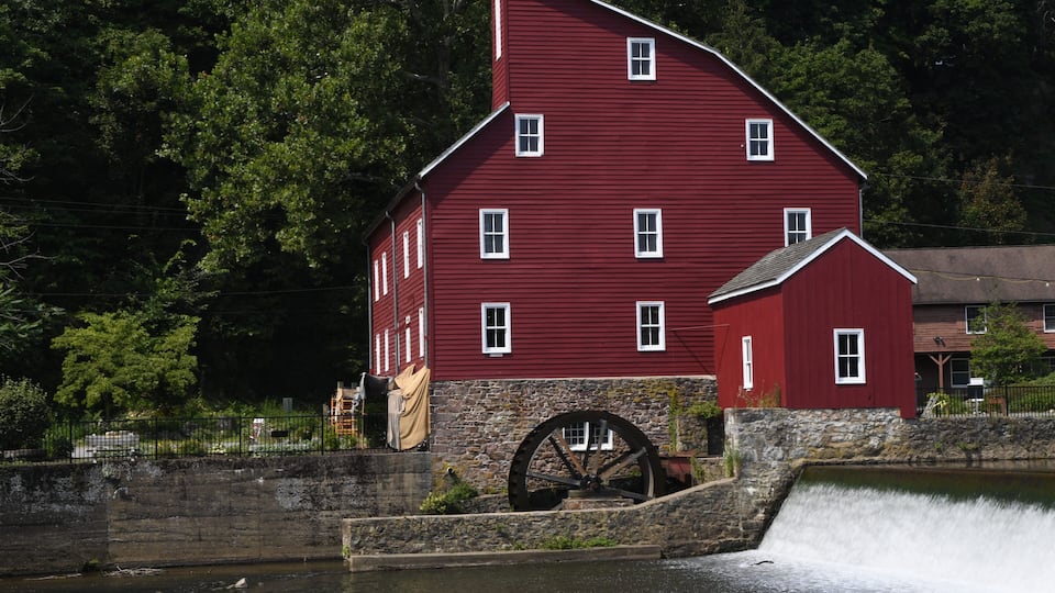 A red watermill with a large wheel, surrounded by a stone wall and lush greenery.
