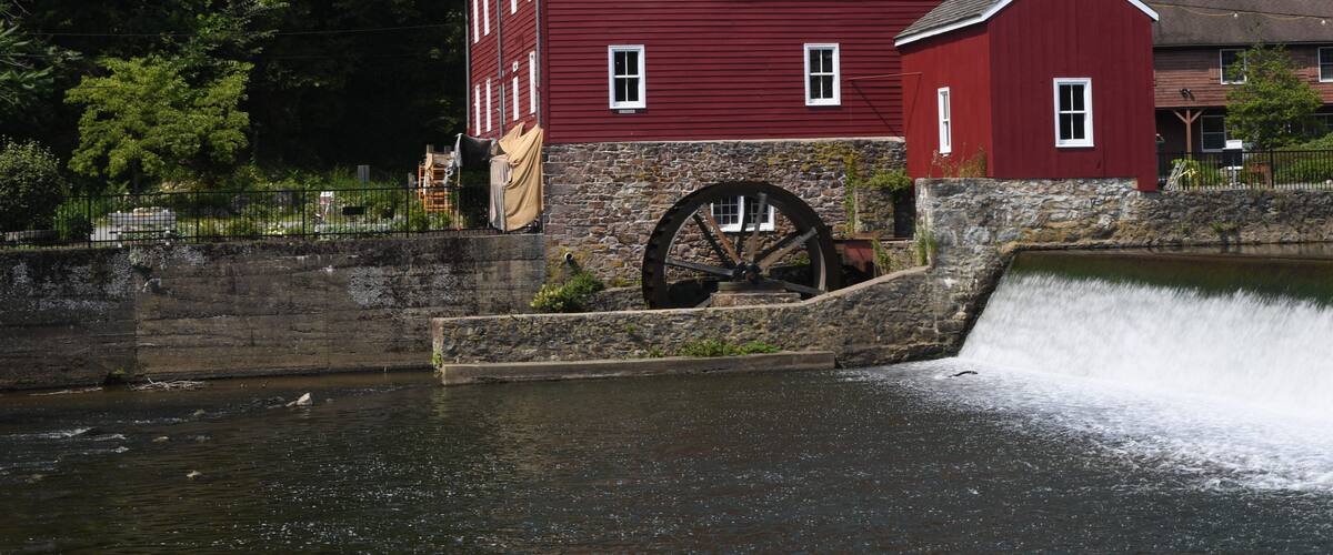 A red watermill with a large wheel, surrounded by a stone wall and lush greenery.