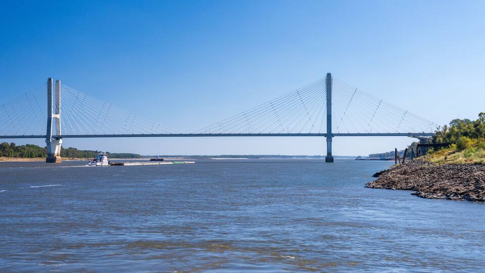 A large suspension bridge over a wide river, with a boat in the foreground and a rocky shoreline on the right.