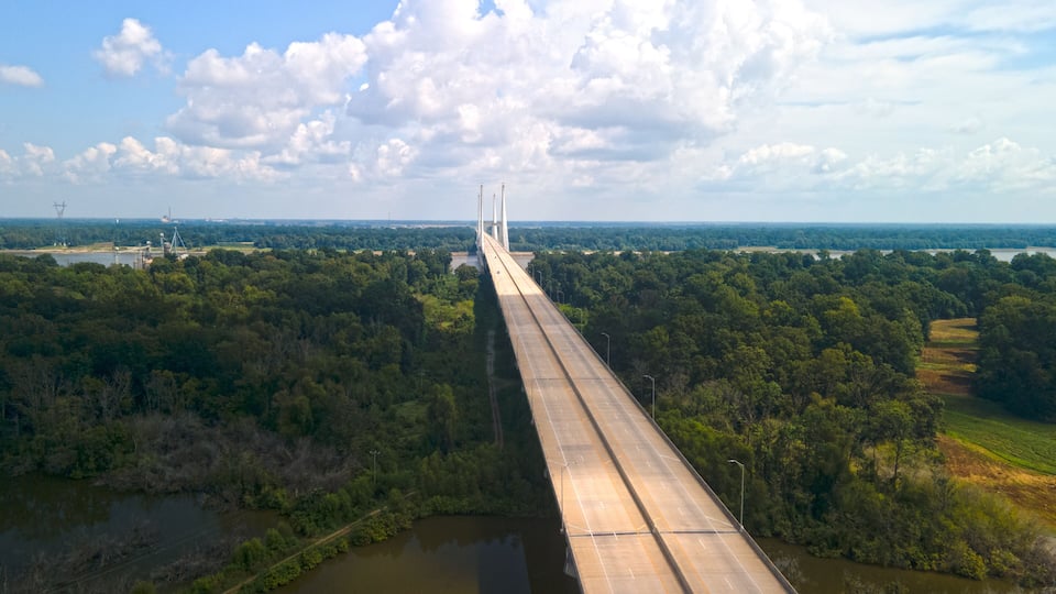 A long bridge spanning a river, surrounded by dense forest and fields.