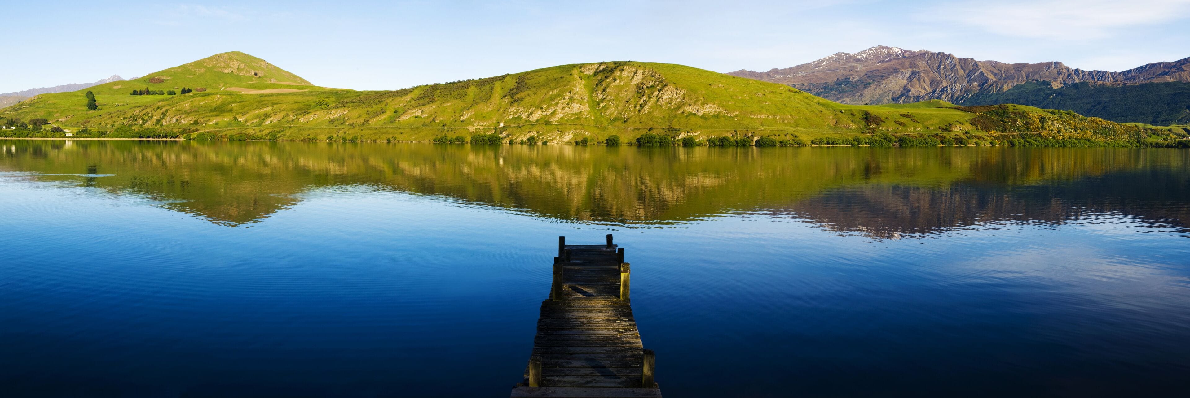 A calm lake with a wooden dock extending into the water, surrounded by green hills and mountains under a clear sky.