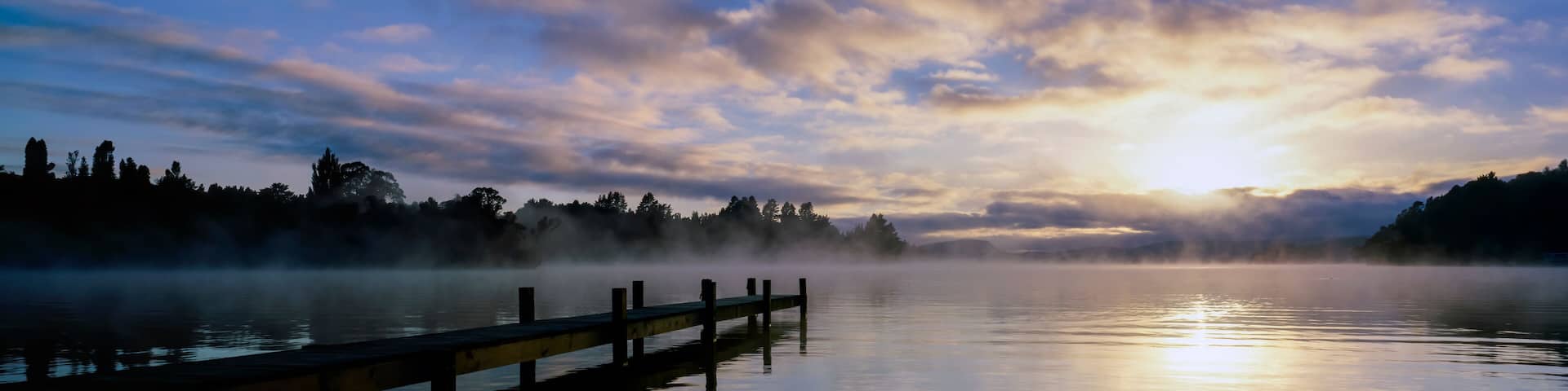 Panorama of mist raising from Lake Rotoiti at sunrise and wooden jetty stretching out from shore