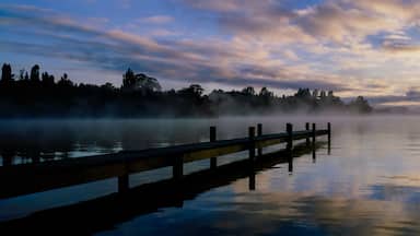 A calm lake at sunrise with a wooden dock extending into the water, surrounded by trees and a misty atmosphere.