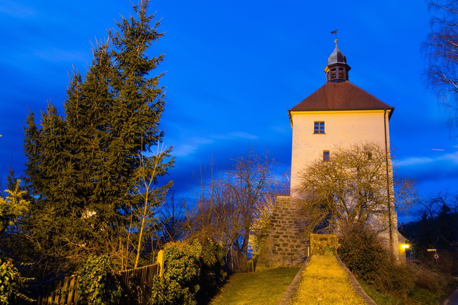 Ein Turm mit rotem Dach und Wetterfahne, umgeben von Bäumen und einer Steinmauer.
