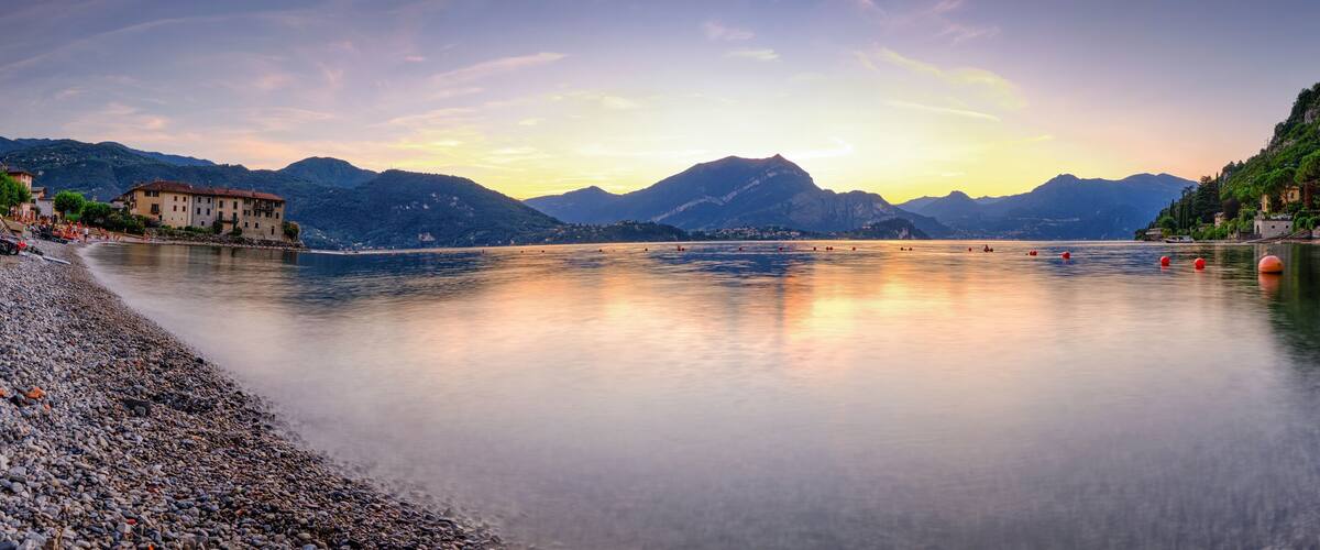 A calm lake with mountains in the background, a pebbled beach, and buildings along the shore.