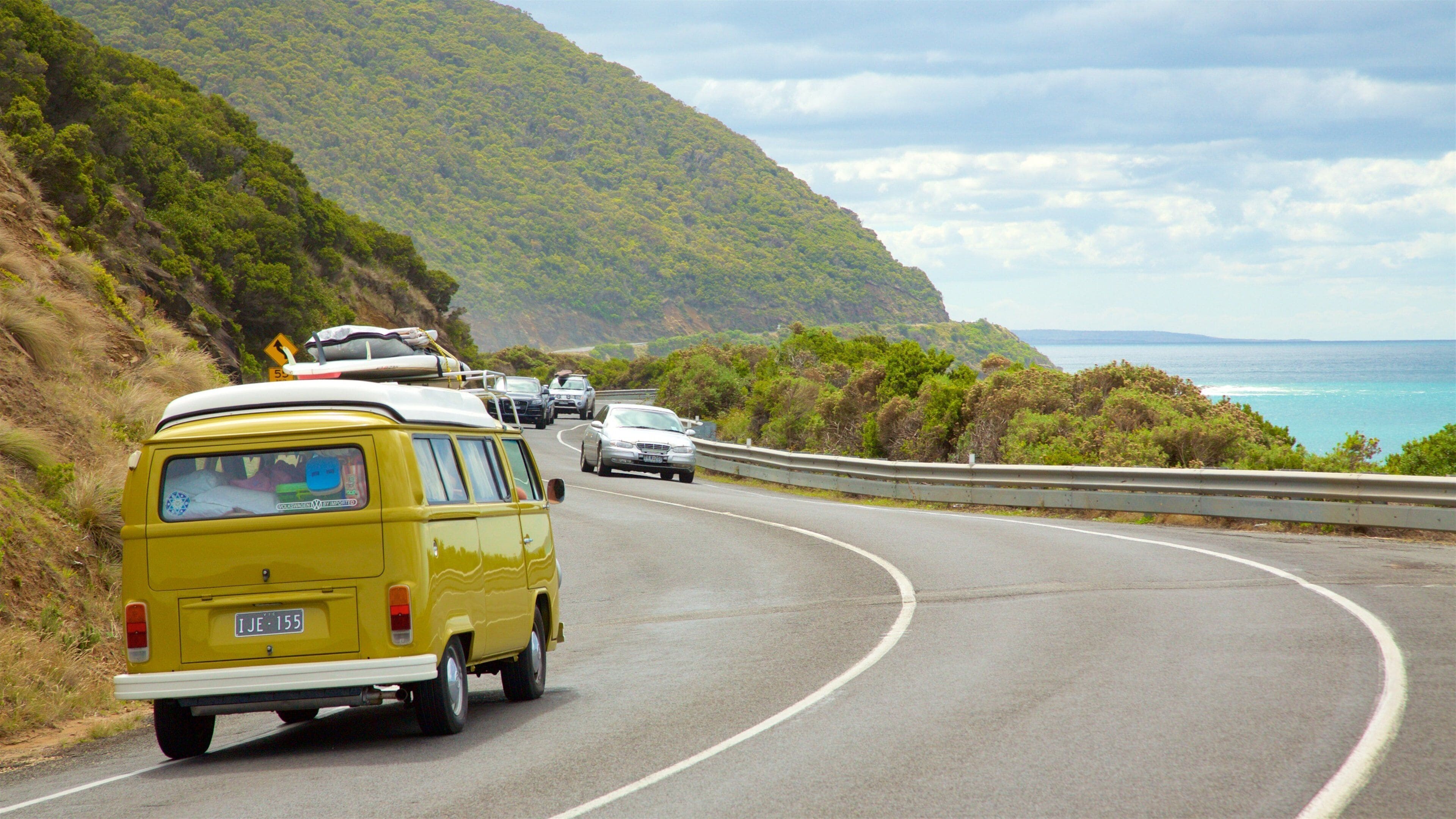 A winding coastal road with a yellow van and several cars.