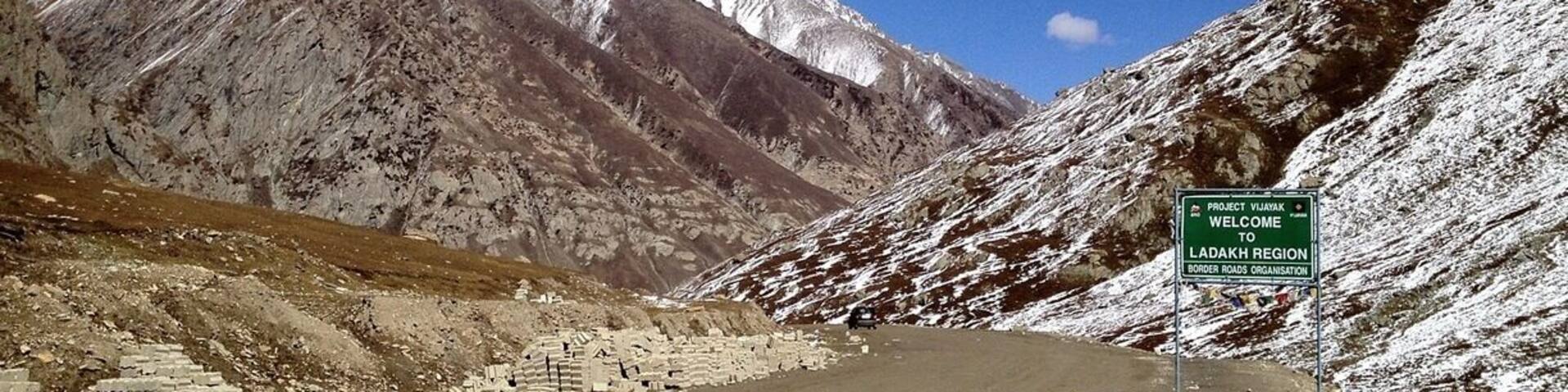 A road leading to a snow-capped mountain range with a sign welcoming visitors to the Ladakh region.