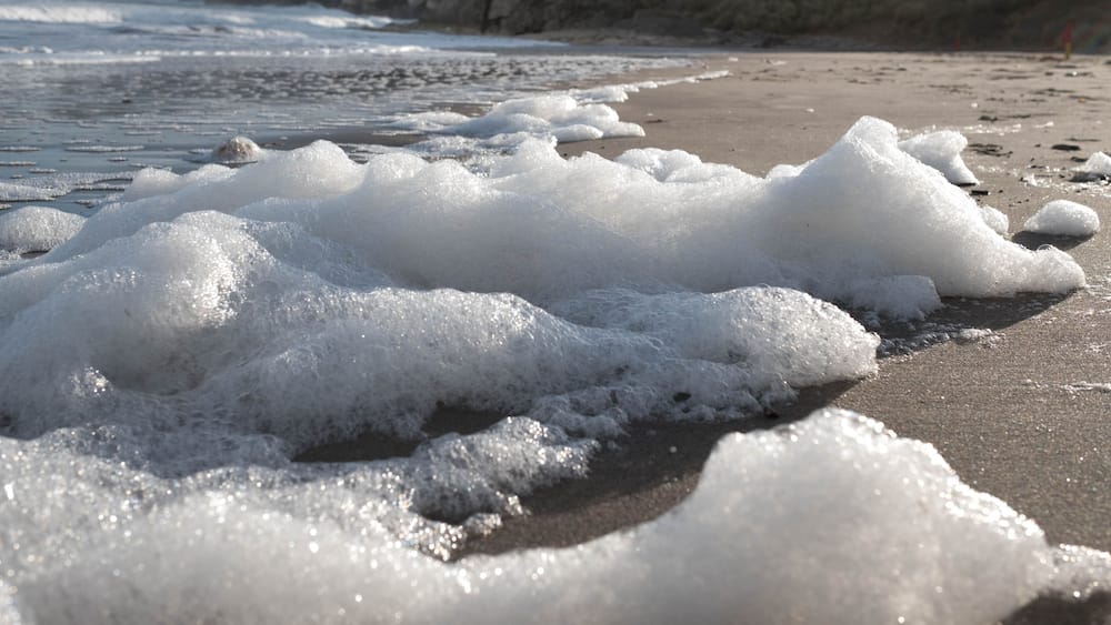 Une plage avec des vagues et de l’écume, des falaises rocheuses en arrière-plan.