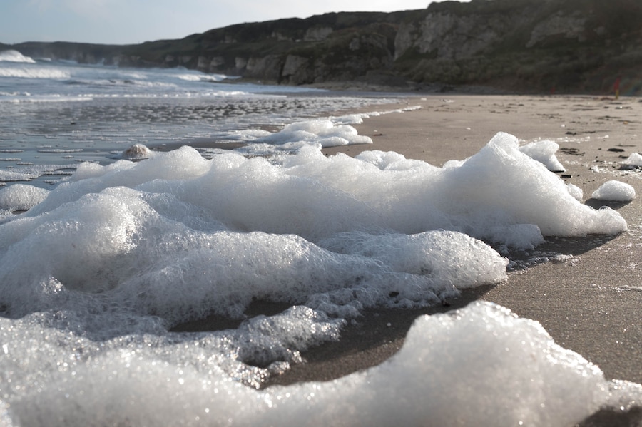A beach with waves and foam, rocky cliffs in the background.