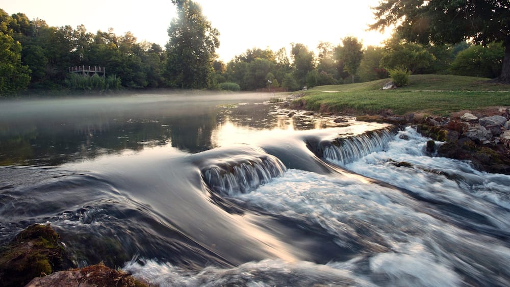 A waterfall cascading over rocks into a calm river surrounded by trees and a grassy area.