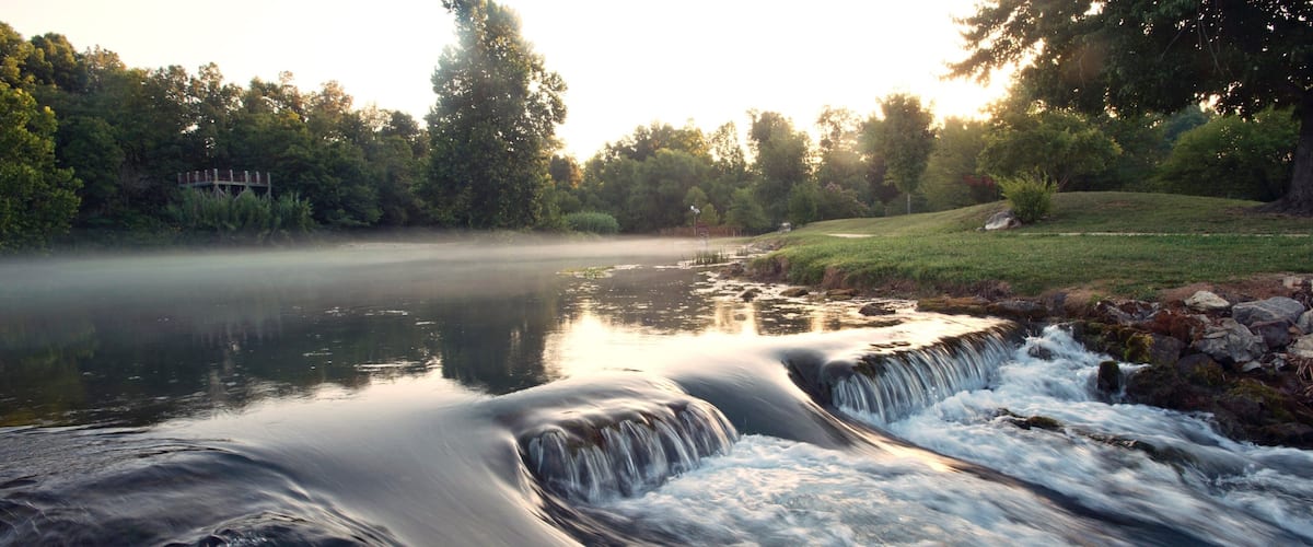 A waterfall cascading over rocks into a calm river surrounded by trees and a grassy area.