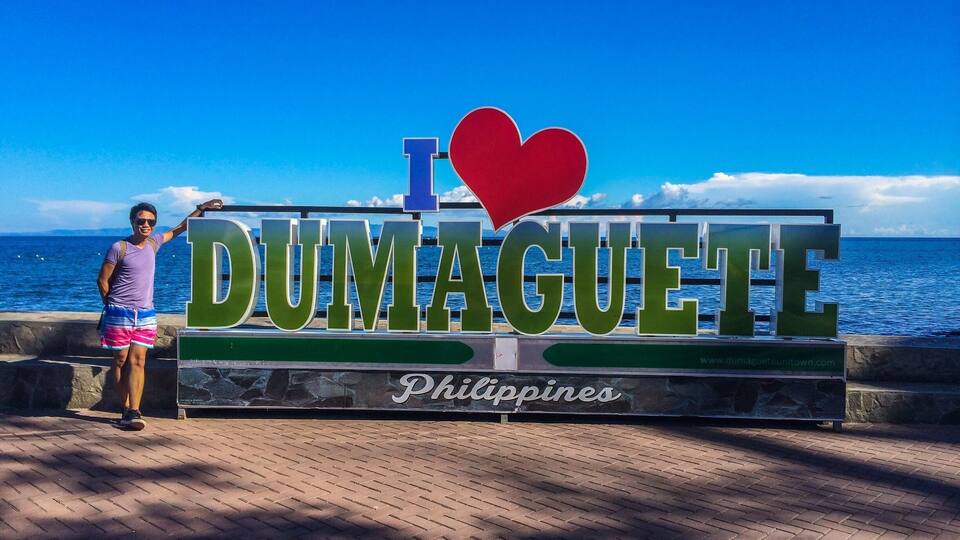 A person posing in front of a sign that reads "I love Dumaguete, Philippines" with a view of the ocean in the background.