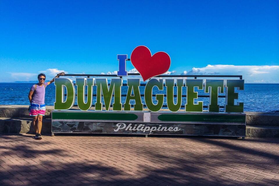 A person posing in front of a sign that reads "I love Dumaguete, Philippines" with a view of the ocean in the background.