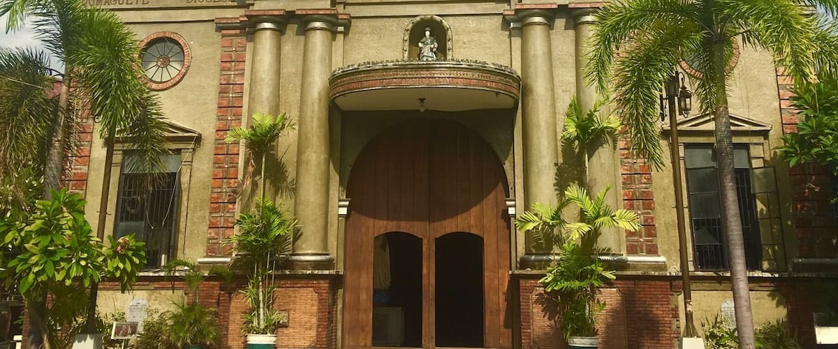 A historic church with a central dome, two statues on the facade, and a large wooden entrance.