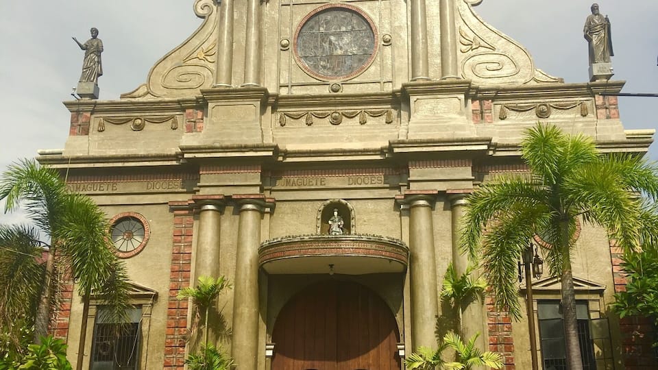 A historic church with a central dome, two statues on the facade, and a large wooden entrance.