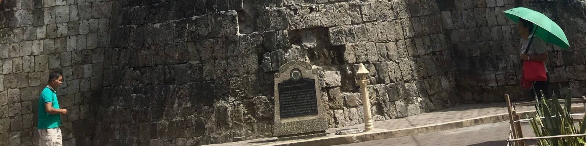 A historic stone tower with a clock and a plaque, surrounded by buildings and greenery.