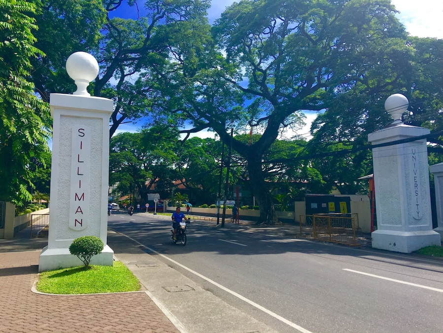 Une rue bordée d’arbres, avec deux monuments blancs portant les inscriptions « SILLIMAN » et « UNIVERSITY », et un ciel dégagé en arrière-plan.