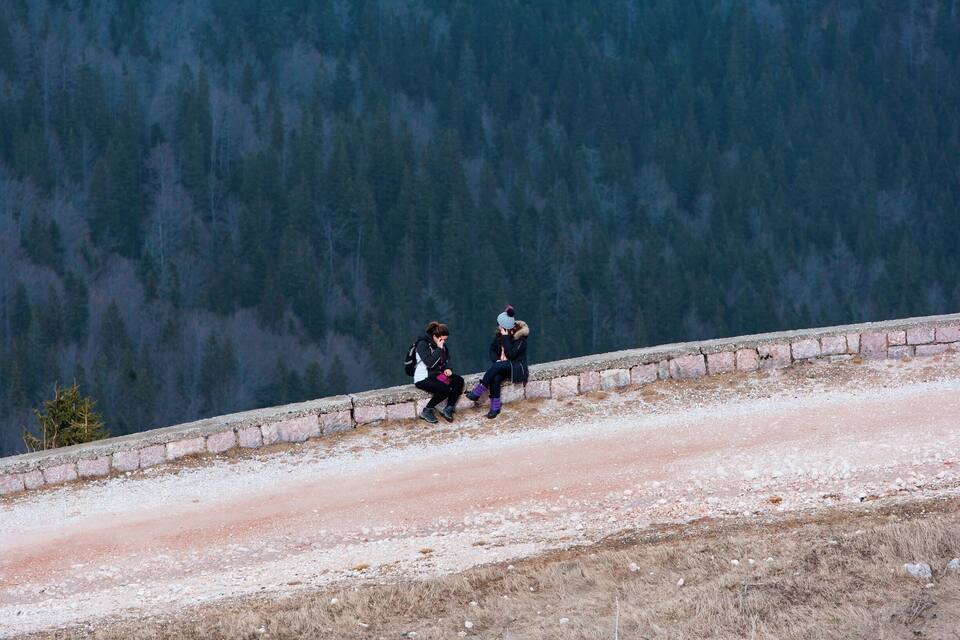 Duas pessoas sentadas em um degrau de pedra com vista para um vale arborizado.