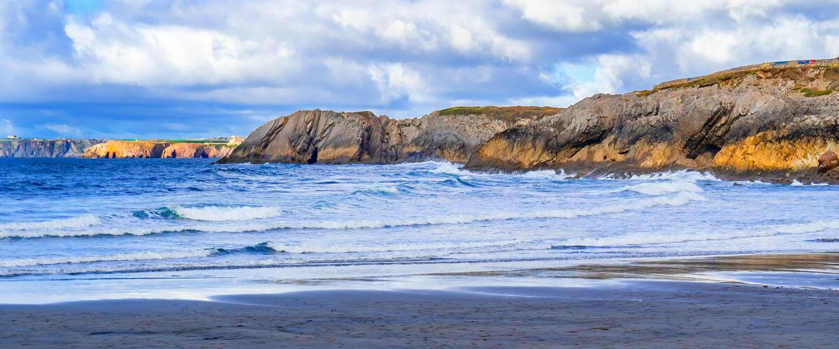 A rocky coastline with waves crashing onto the shore, a clear sky, and distant buildings.