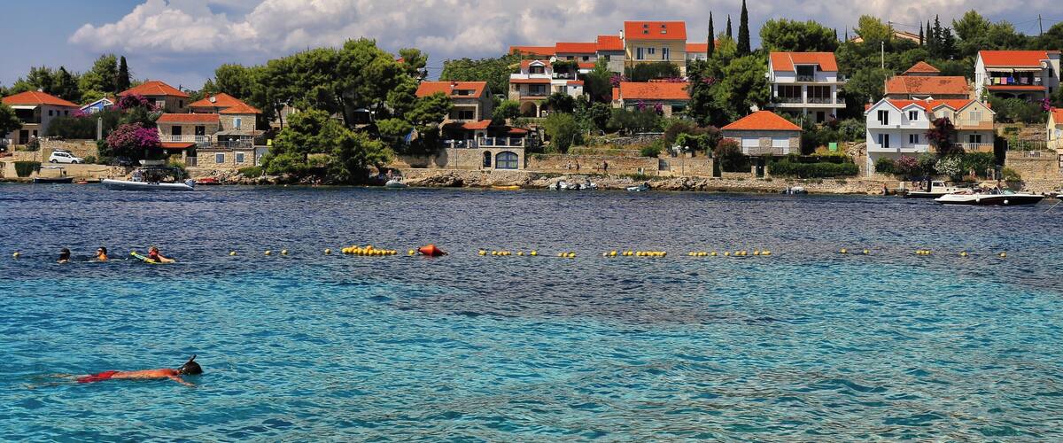 A coastal town with red-roofed buildings, clear blue water, and swimmers.