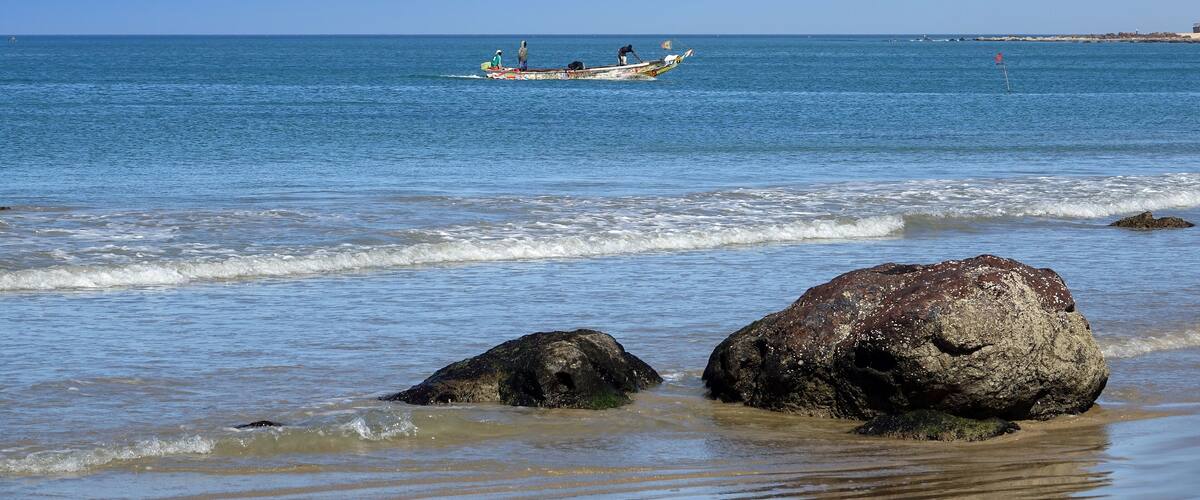 A calm sea with a boat carrying people, large rocks on the shore, and a clear blue sky.