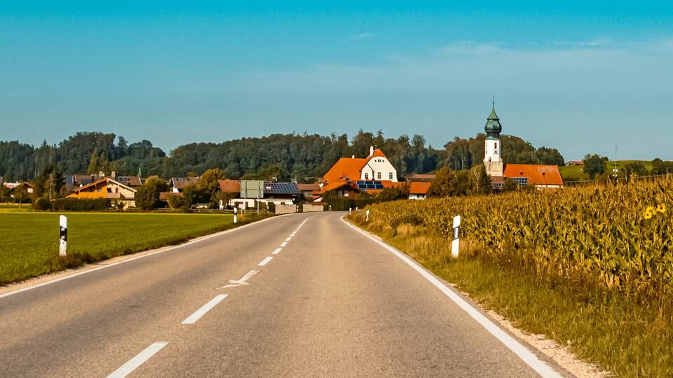 A rural road with a church and a tower in the distance, surrounded by fields and houses.