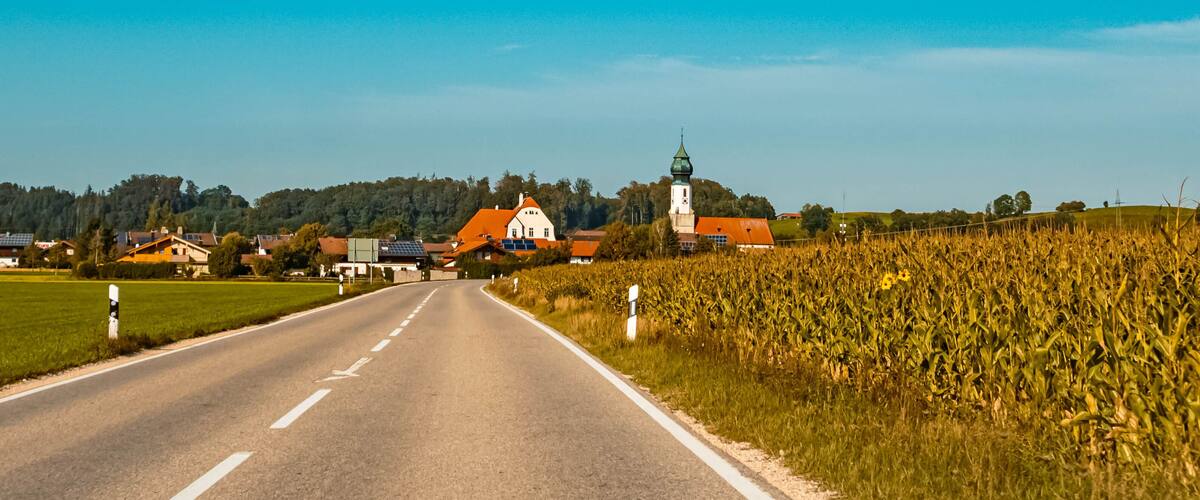 A rural road with a church and a tower in the distance, surrounded by fields and houses.