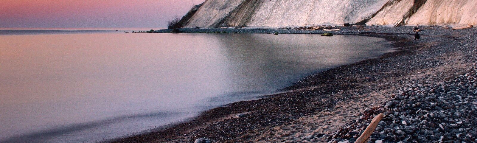 A pebbled beach with a large fallen tree trunk, calm water, and a rocky cliff in the background.