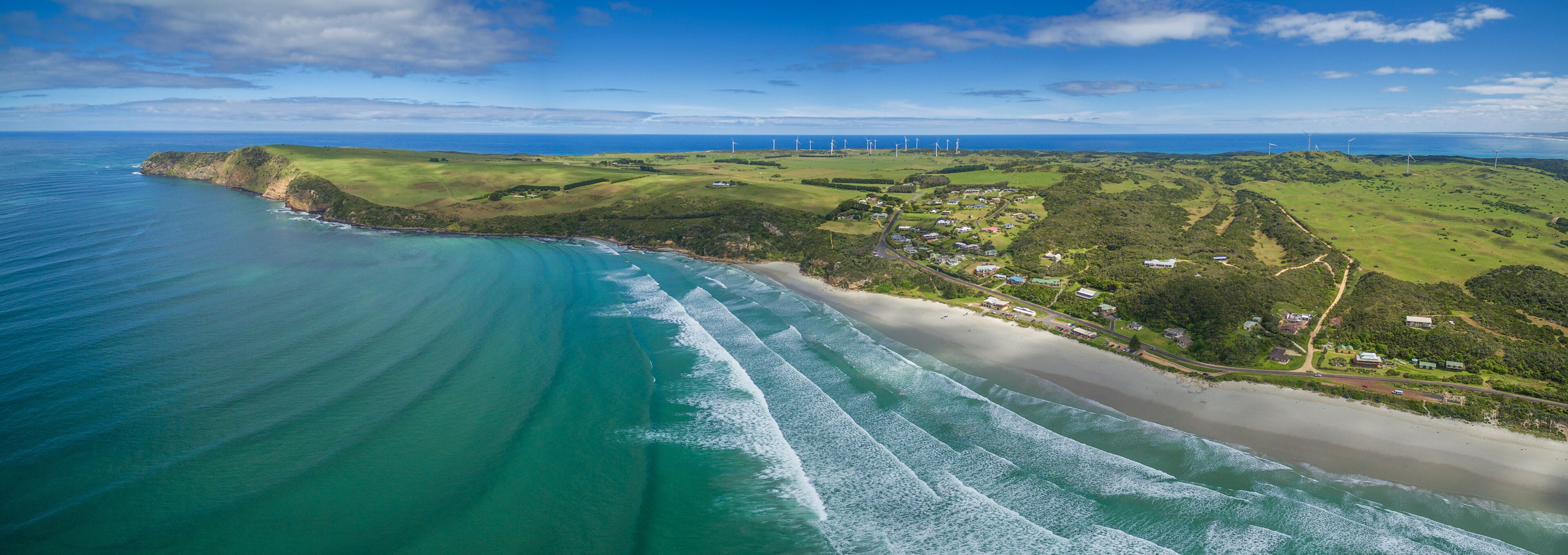 A coastal landscape with a beach, ocean waves, green hills, and wind turbines.