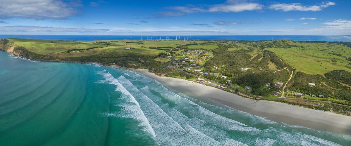 A coastal landscape with a beach, ocean waves, green hills, and wind turbines.
