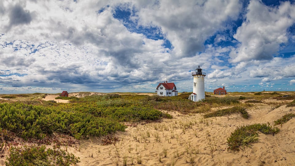 A lighthouse, a red house, and a white house on a sandy beach with dunes and vegetation.