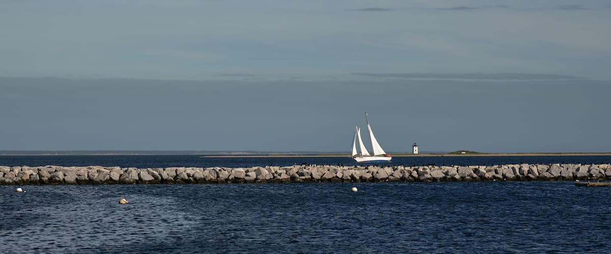 A sailboat on a calm sea, a rocky breakwater, and a lighthouse in the distance.