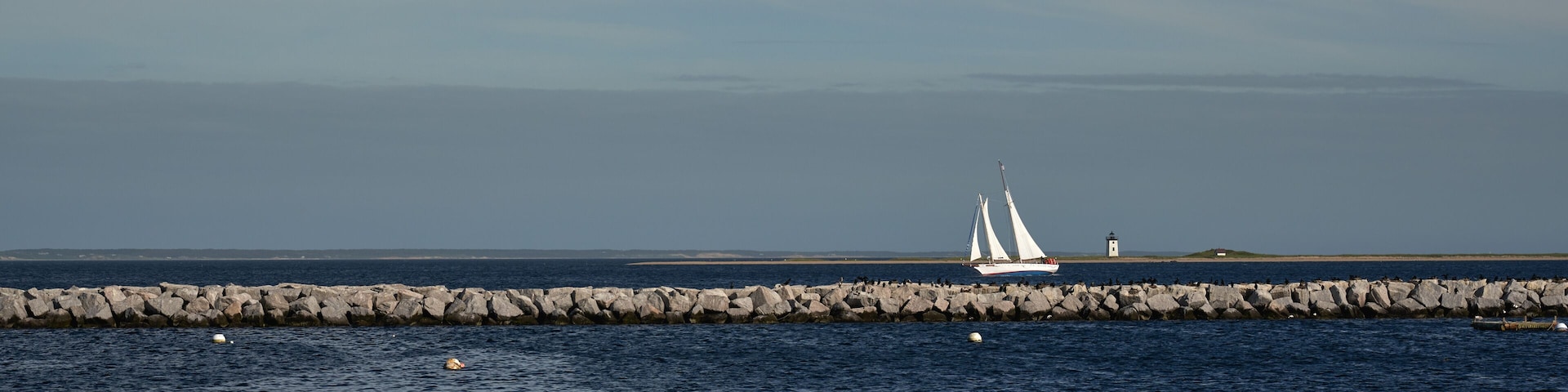 A sailboat on a calm sea, a rocky breakwater, and a lighthouse in the distance.