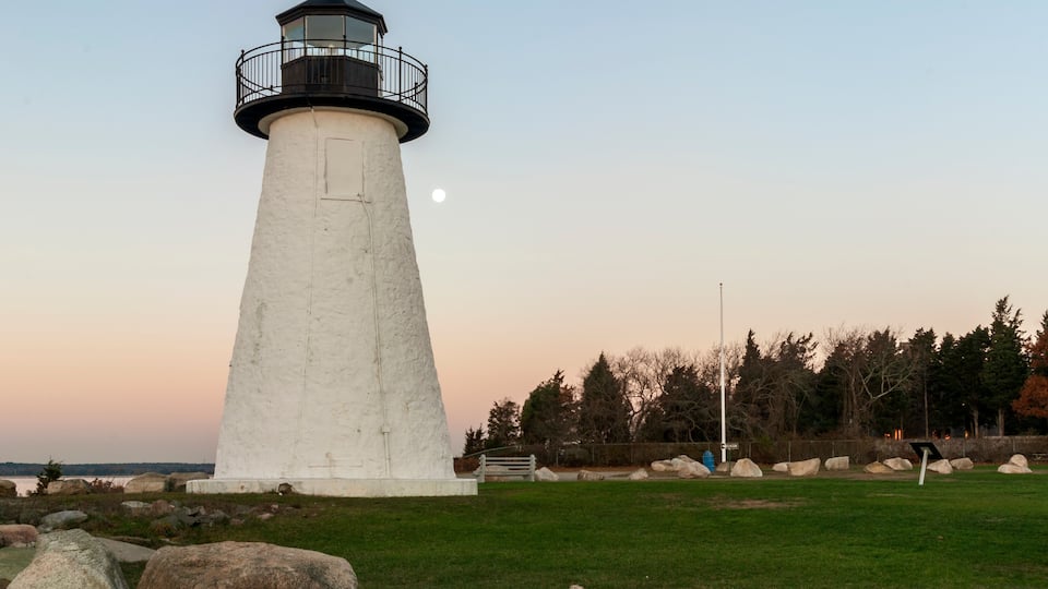 A white lighthouse with a black top, surrounded by rocks and grass, with trees and a bench in the background.
