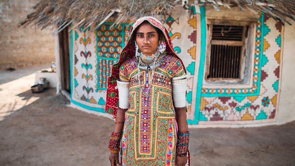 A person in traditional colorful attire standing in front of a decorated house with a thatched roof.
