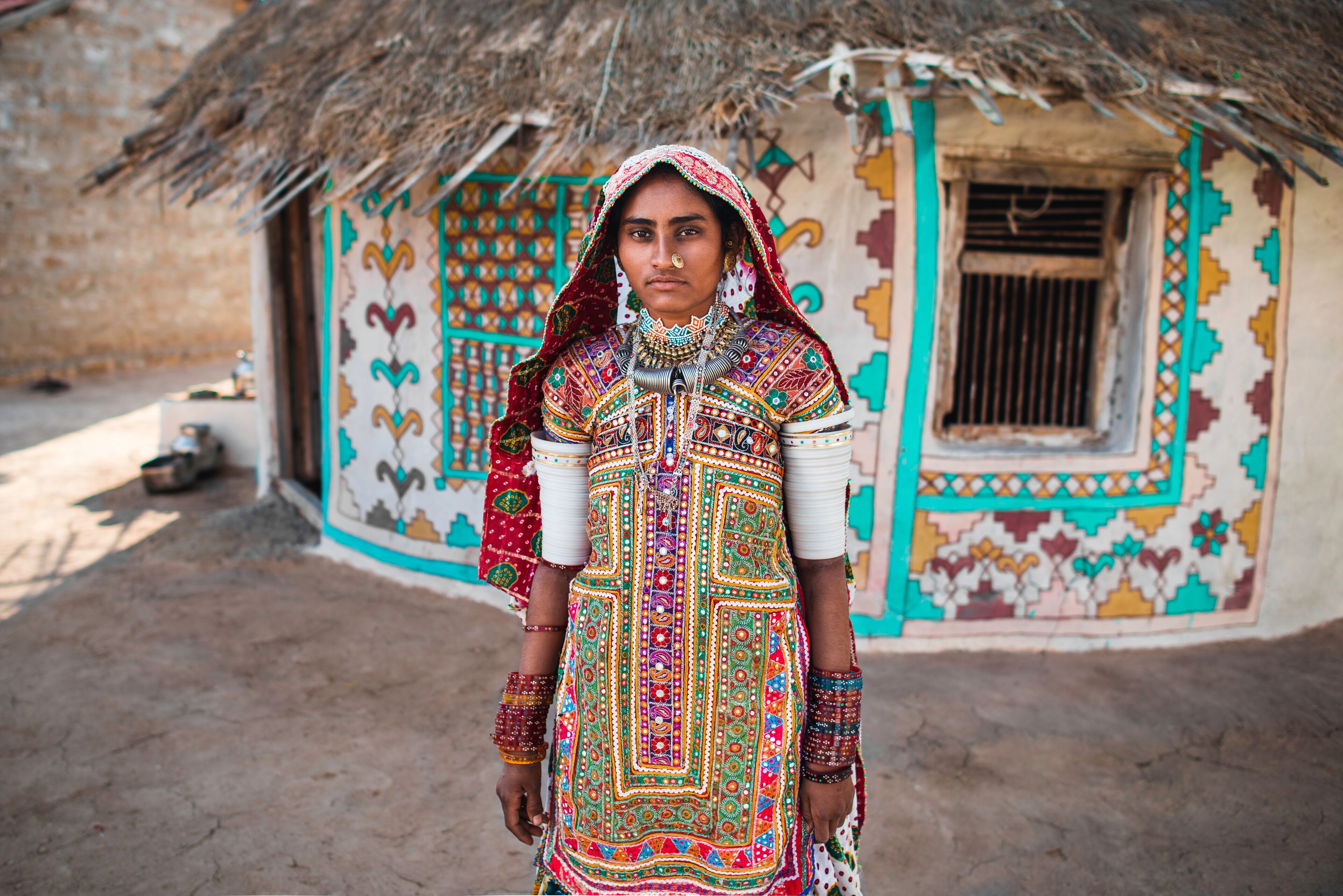 A person in traditional colorful attire standing in front of a decorated house with a thatched roof.