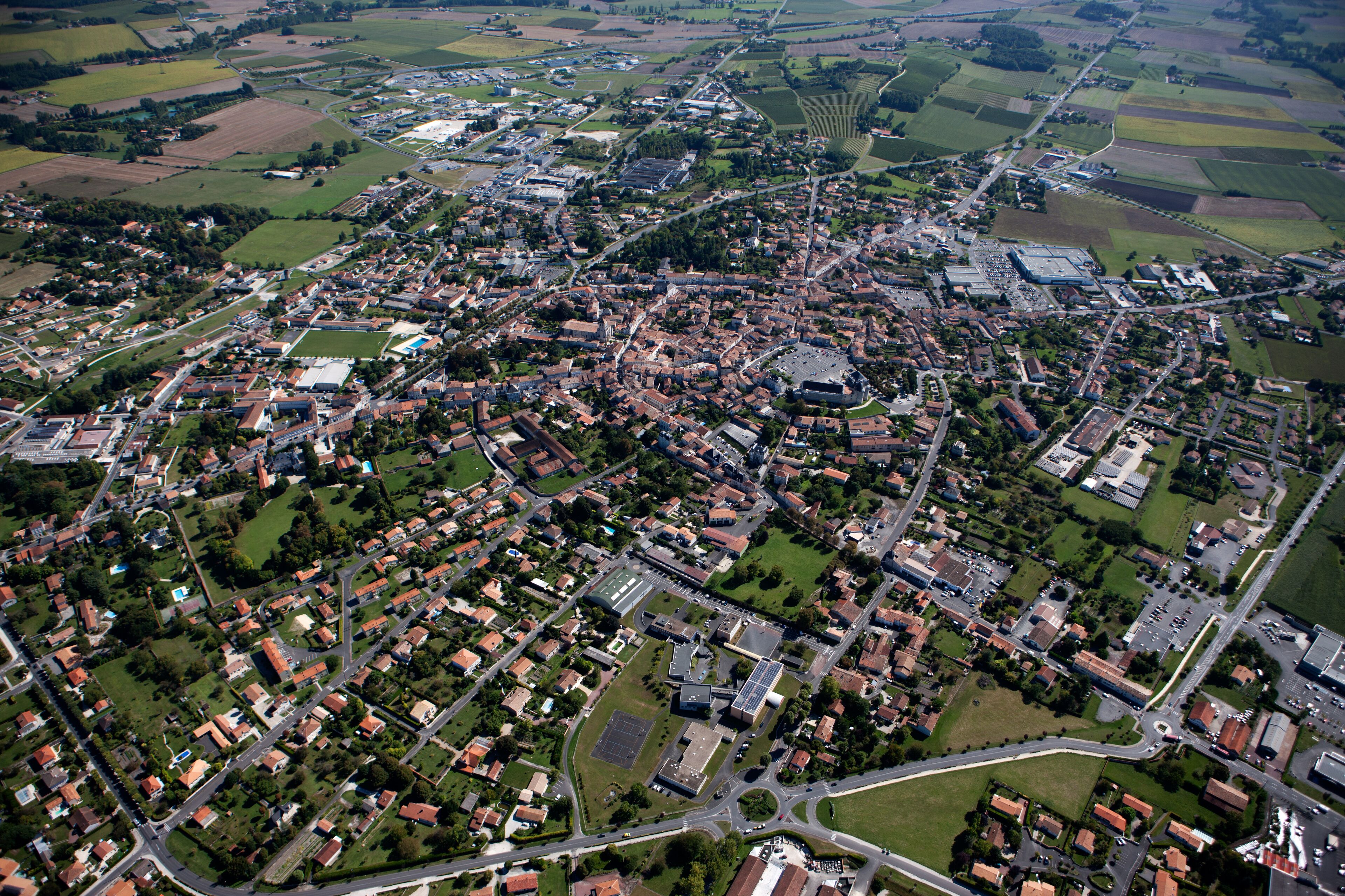 An aerial view of a town with a mix of residential and commercial buildings, surrounded by green fields and roads.